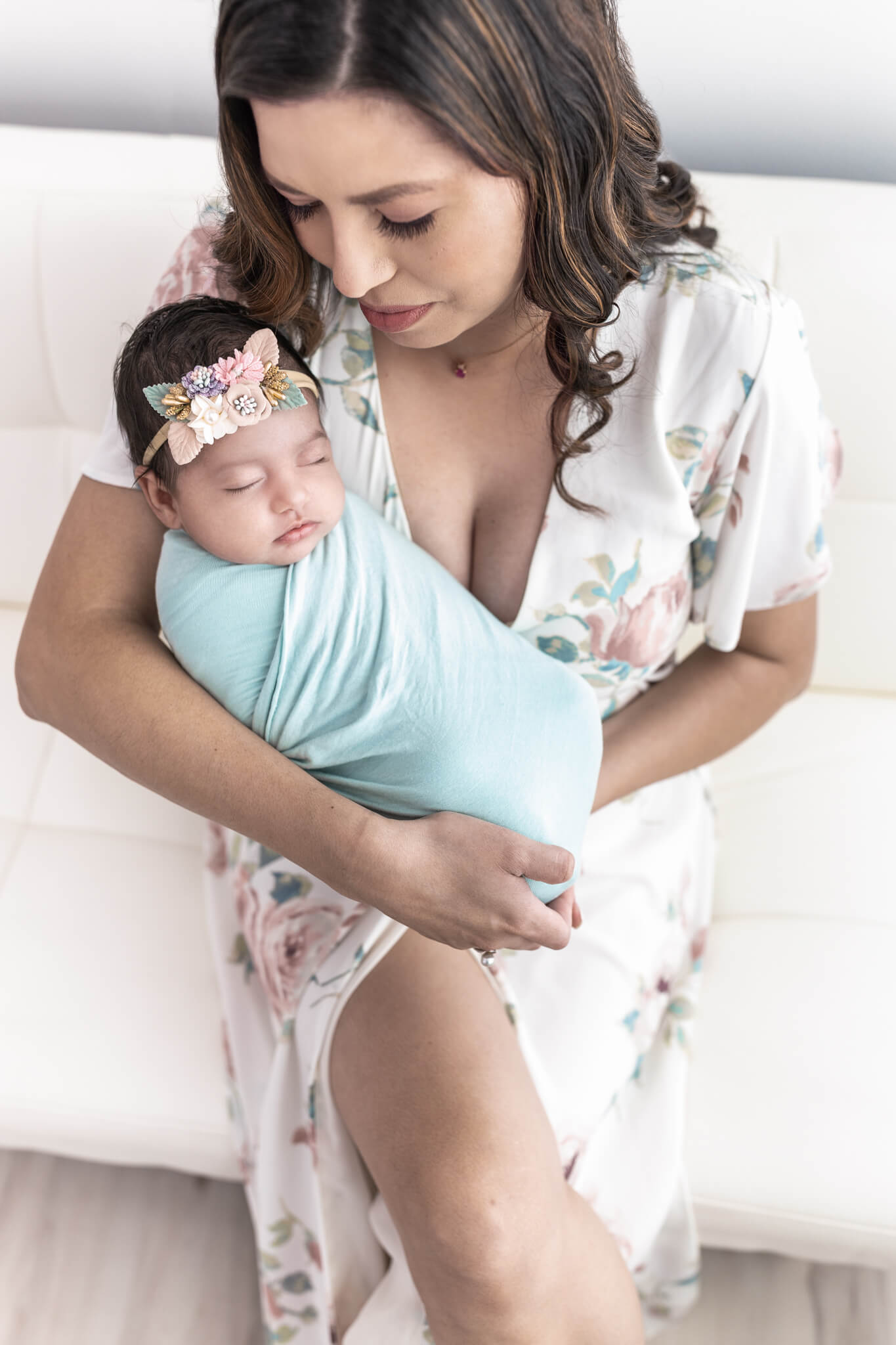 Mom in a cream floral dress sits on a white couch looking down lovingly at her sleeping baby girl during a newborn session.