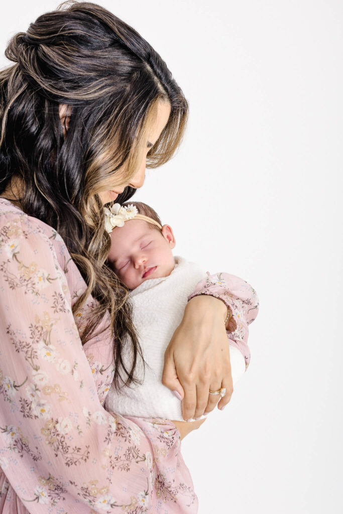 A woman with long wavy hair in a pink floral dress gently cradles a sleeping baby dressed in white with a flower headband, both facing sideways against a plain white background.