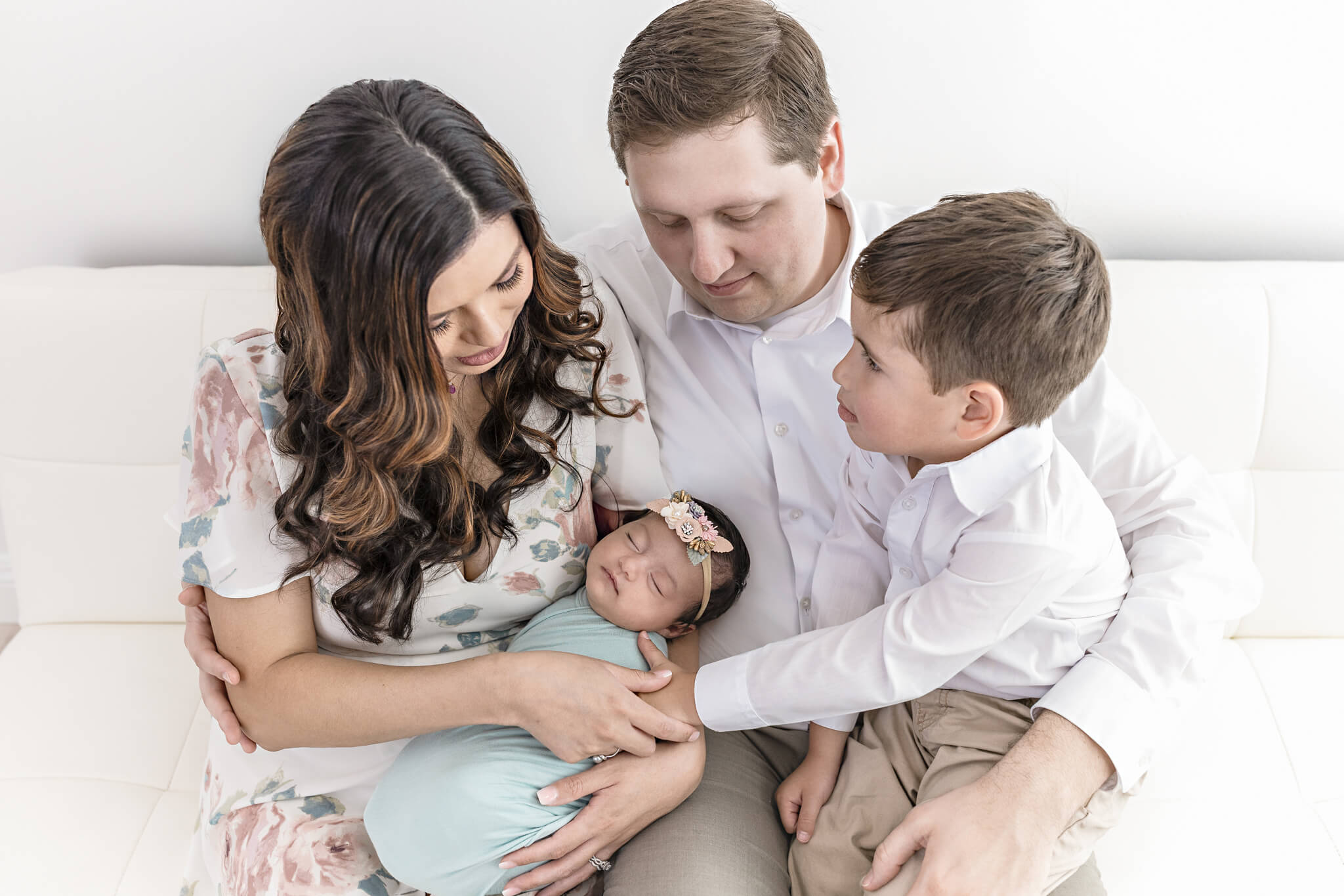 A family of four sits closely on a white couch. The mother holds a swaddled newborn, while the father and young boy sit beside them, all looking lovingly at the baby.
