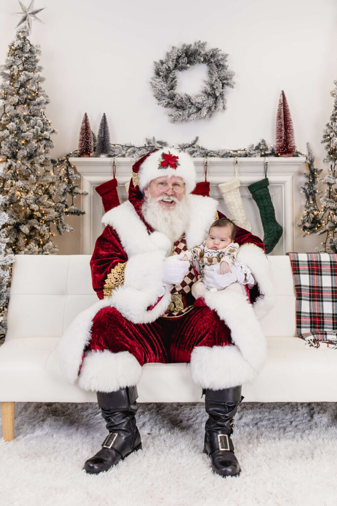 Santa Claus dressed in a red velvet suit sits on a white couch holding a baby. Behind them are decorated Christmas trees, a white mantel with stockings, and a festive wreath on the wall.