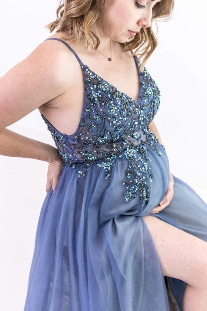 A pregnant woman in a sheer, blue, beaded dress gently cradles her belly and looks down, with her other hand resting on her lower back.
