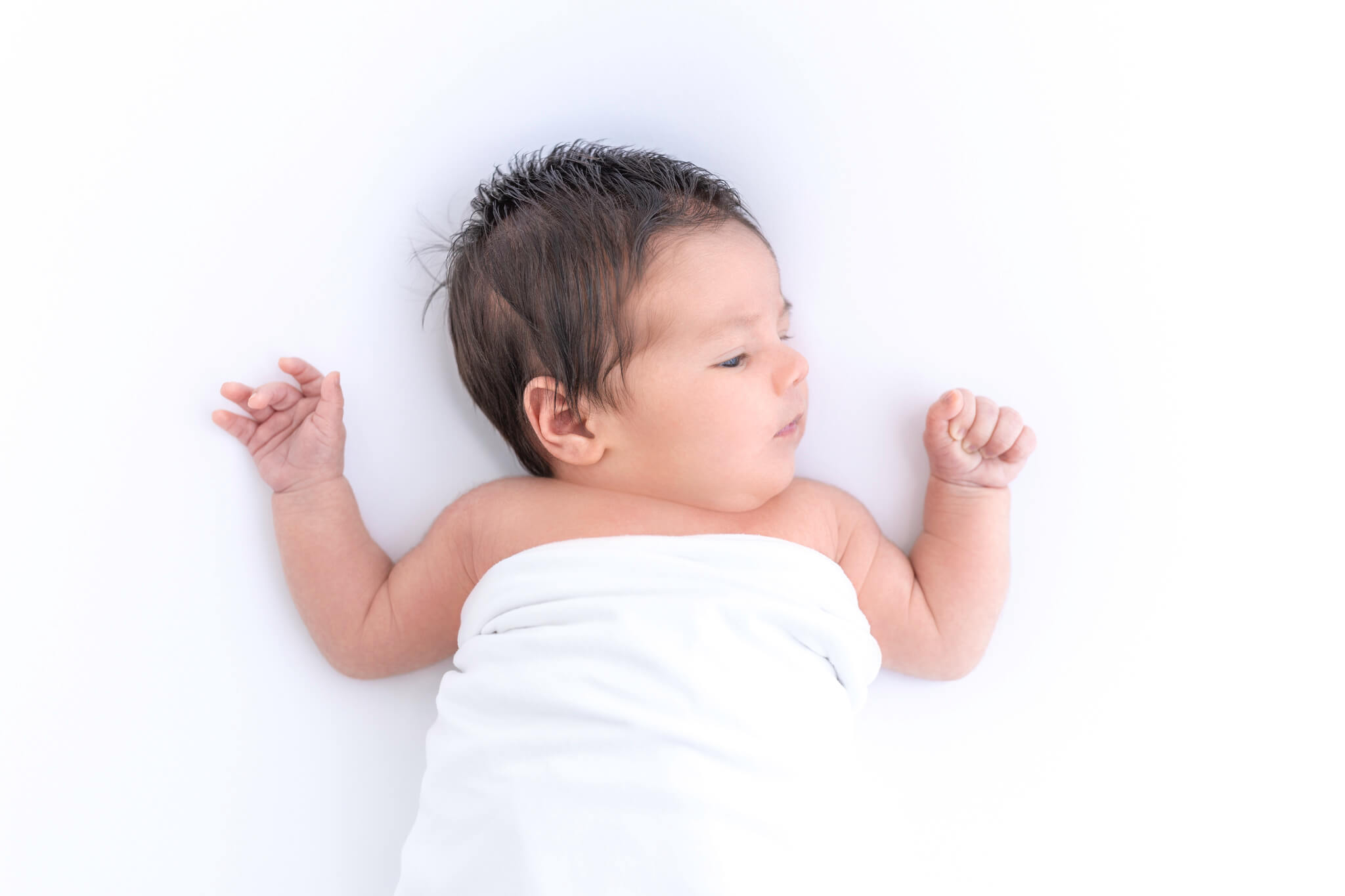 A newborn baby with dark hair lies on a white fabric wrapped in a white blanket looking to the right with arms slightly raised.