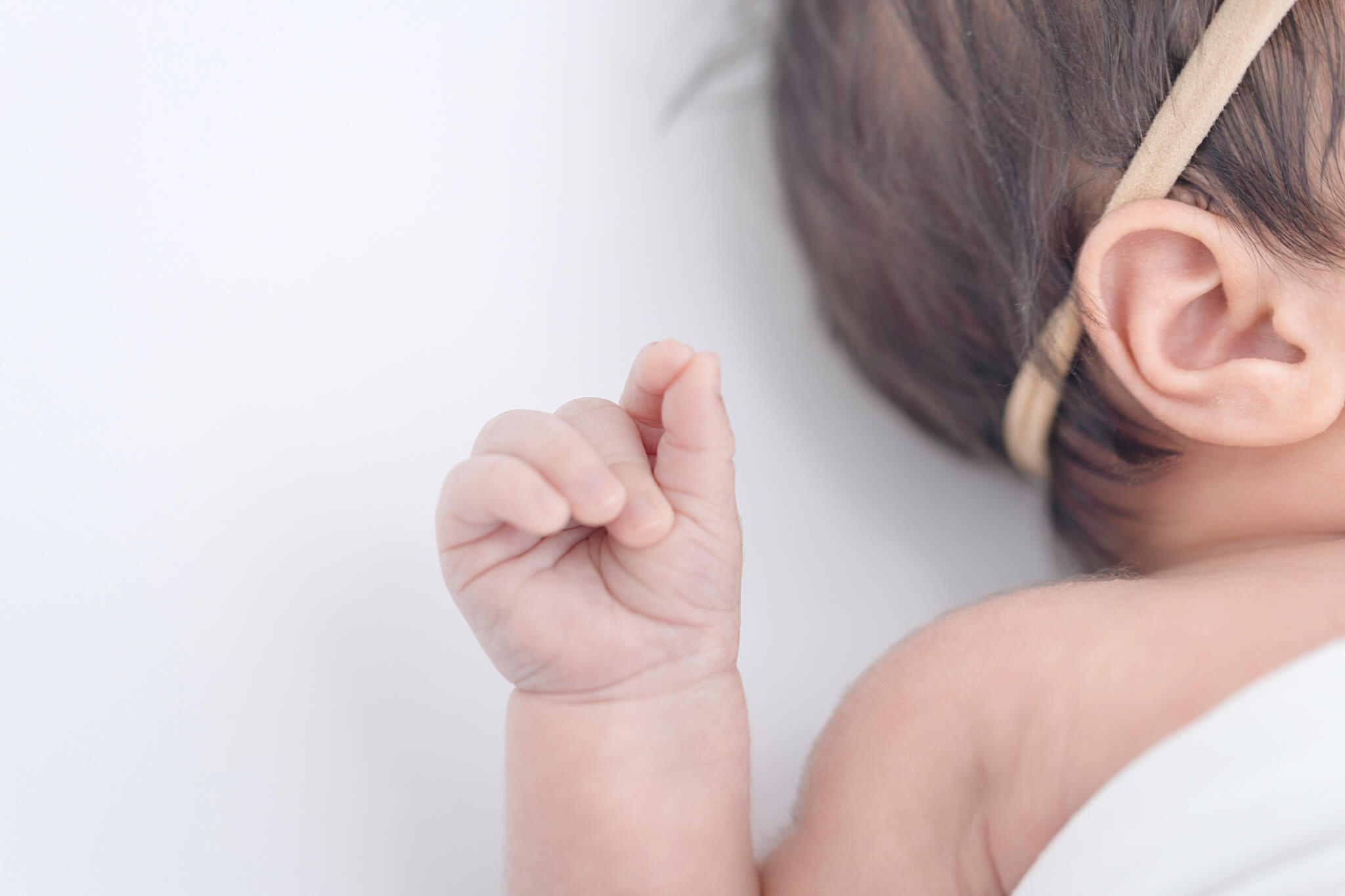 A newborn baby's tiny curled fist beside her ear and headband on a soft white backdrop.