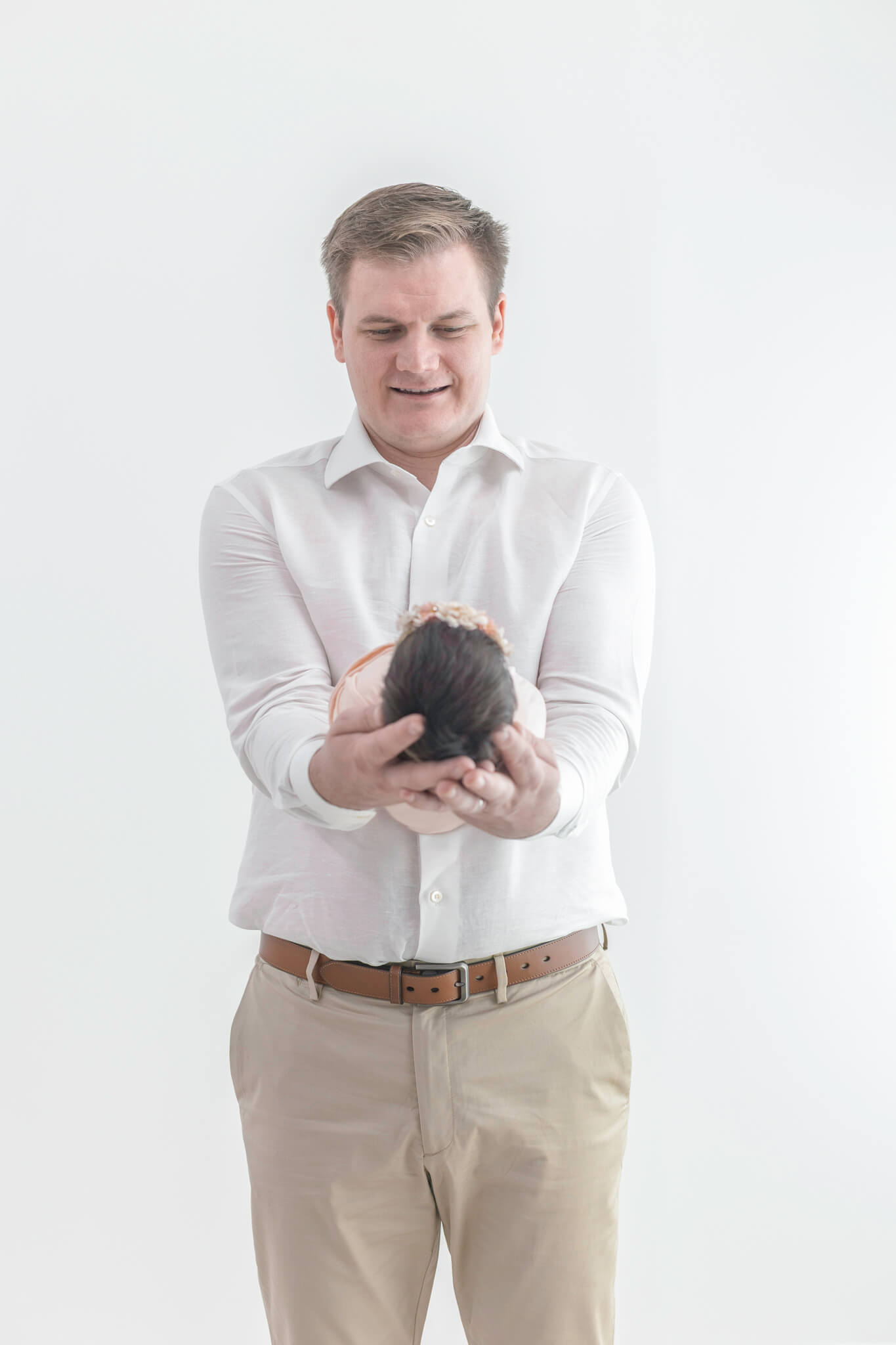 Dad in a white shirt and khakis stands smiling down at his newborn daughter with a floral headband cradled in his hands.
