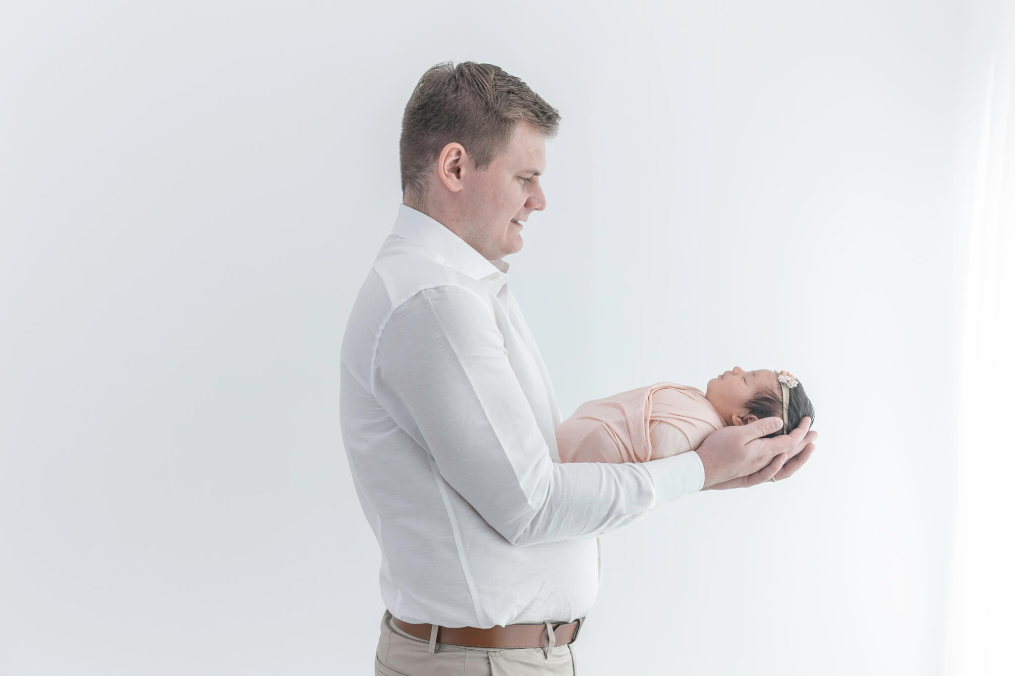A dad in a white shirt gently holds his swaddled newborn baby in his arms, standing against a bright white background.