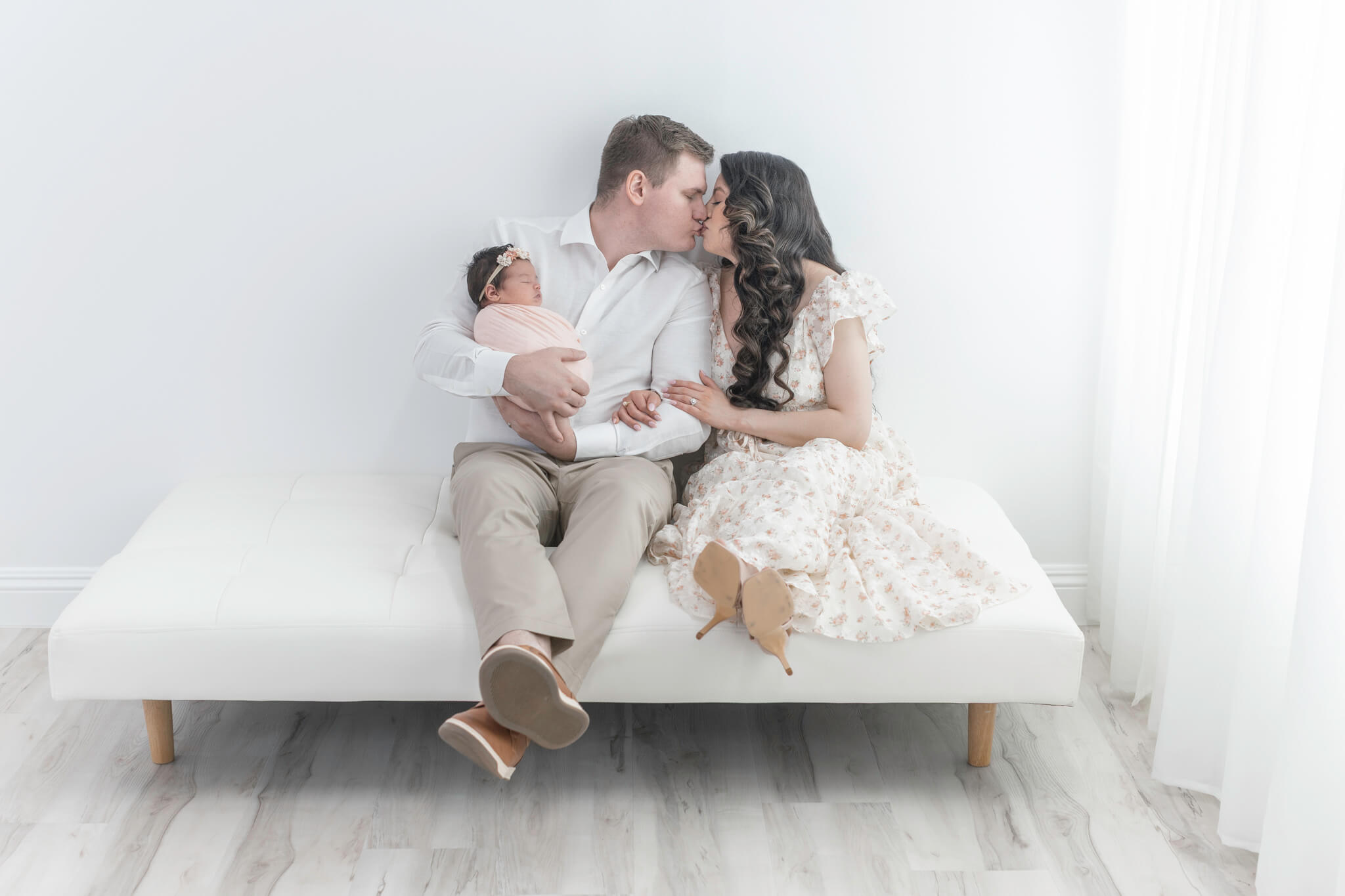 new parents kissing while dad holds new baby girl in peach swaddle and mom slips her arms through his.