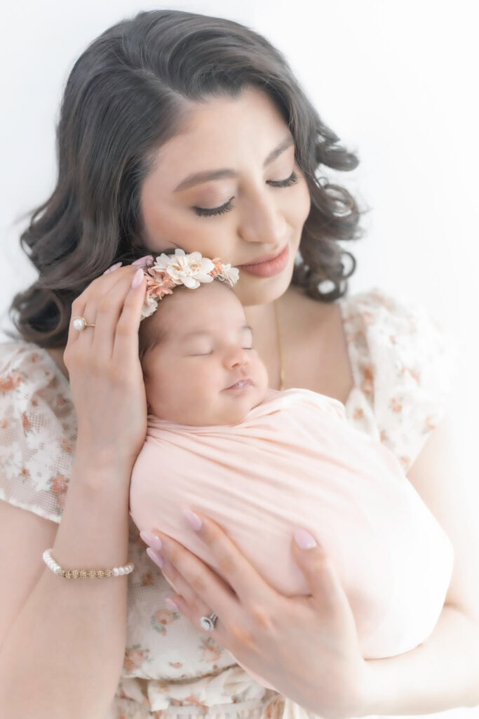 A woman with wavy brown hair lovingly holds a sleeping newborn wrapped in a pale pink blanket. The baby wears a flower headband, and the woman gently cradles the baby’s head, both appearing peaceful.