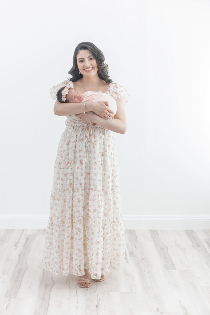 A woman in a long, light-colored floral dress stands barefoot on a light wood floor, smiling and holding a newborn baby in her arms against a bright, plain background.