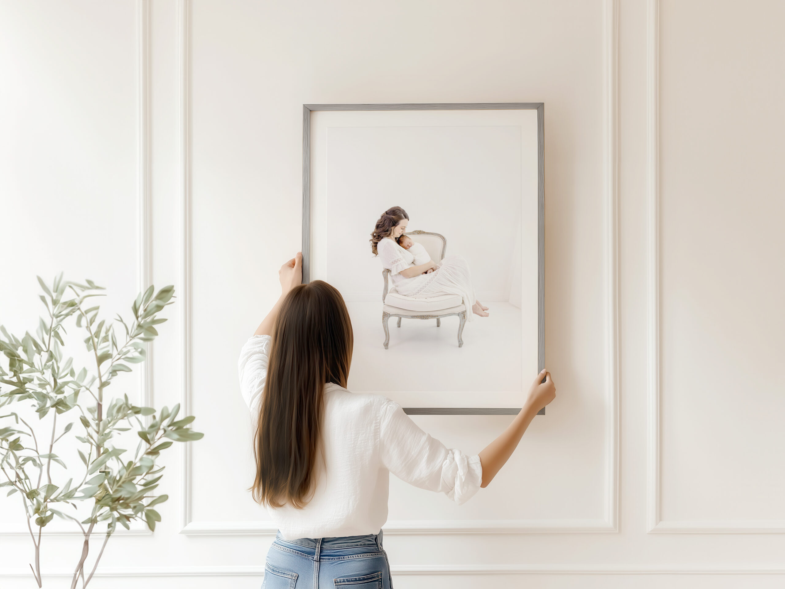 A woman with long brown hair, wearing a white top and jeans, hangs a framed painting from a luxury portrait experince of a seated person on a white wall in a bright, modern room with a green leafy plant nearby.