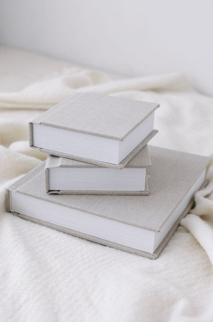 A stack of three hardcover books with plain, light gray covers sits on a soft, cream-colored blanket. The books are arranged in a slightly uneven pile.