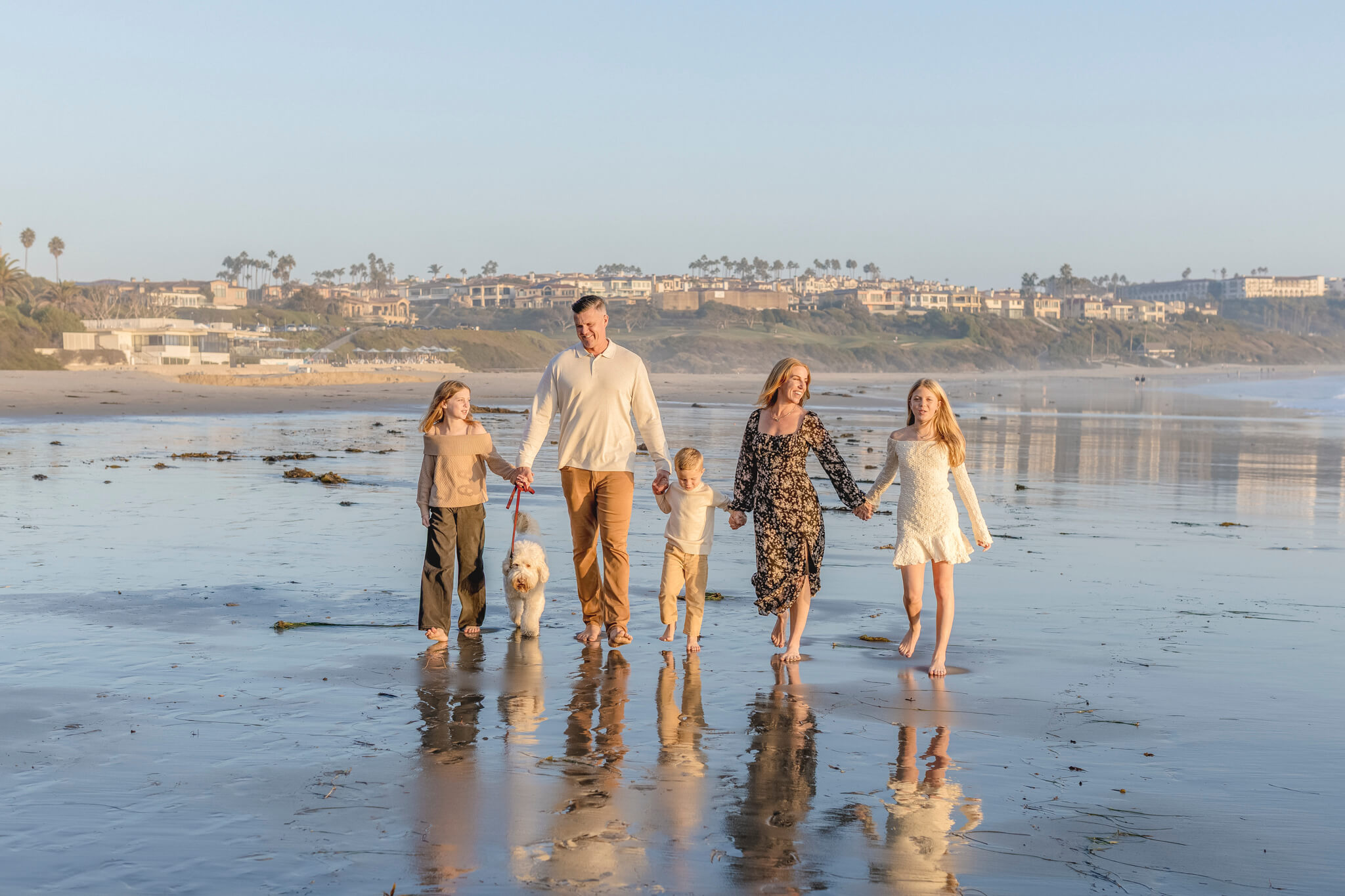 A family of five, including two adults, three children, and a dog, walk hand in hand along a sandy beach at sunset, one of our favorite family photo poses.