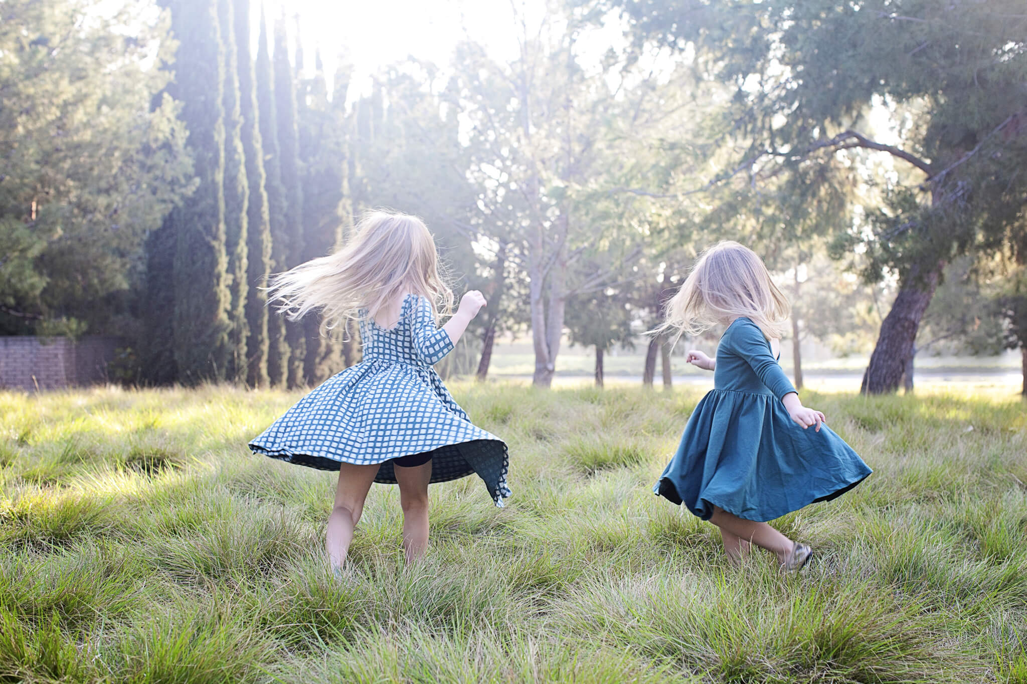 Two young girls in dresses spin and play in a grassy field surrounded by trees.
