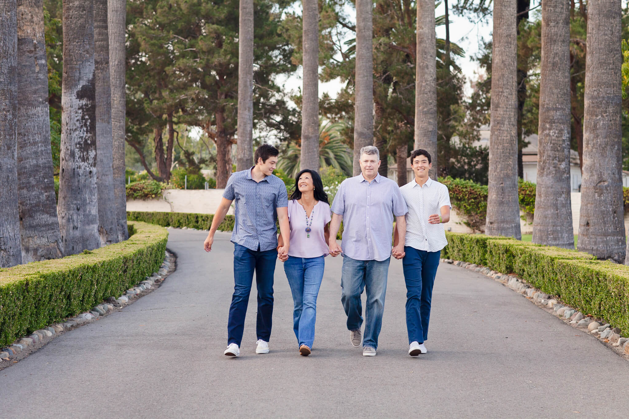 A family of four, dressed in casual clothes, walks together down a tree-lined path. They are smiling and holding hands.