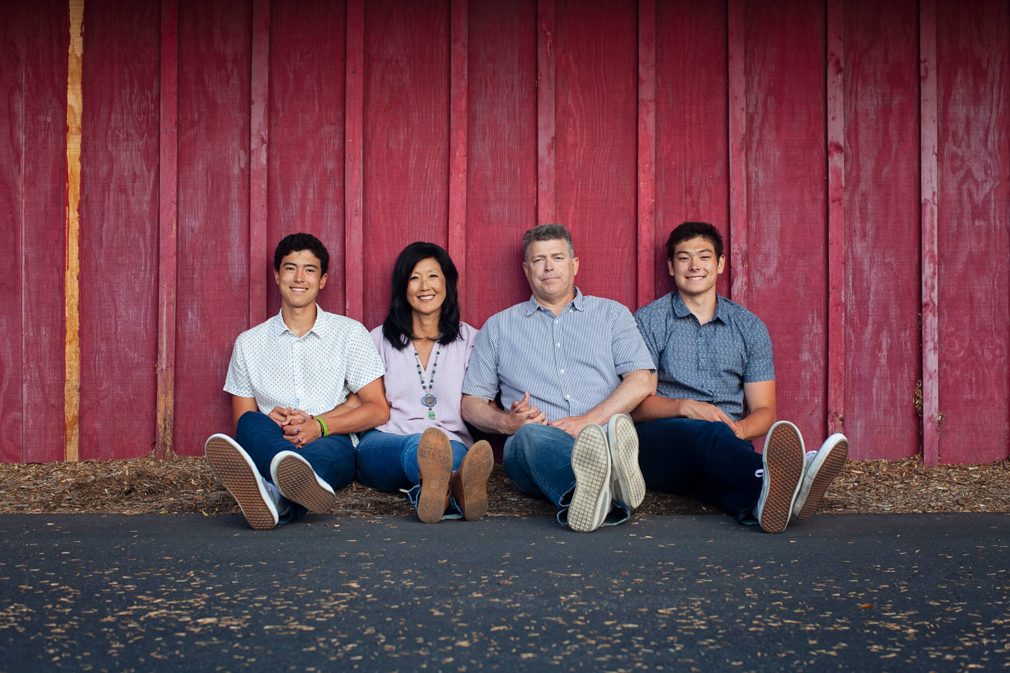 A family of four sits on the ground in front of a red wooden wall, smiling. 