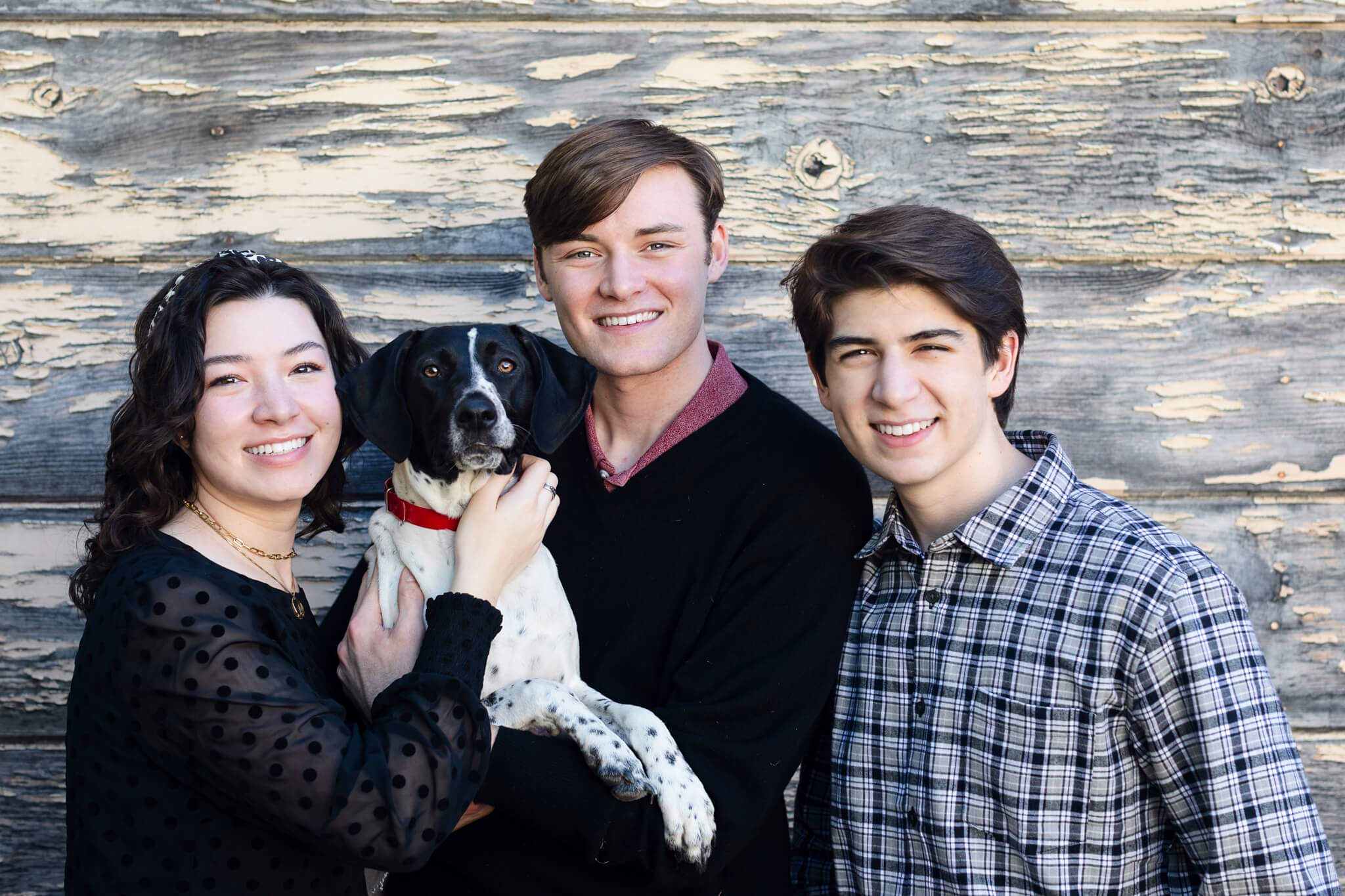 Three young adults smile at the camera while standing in front of a wooden wall holding a black and white dog.