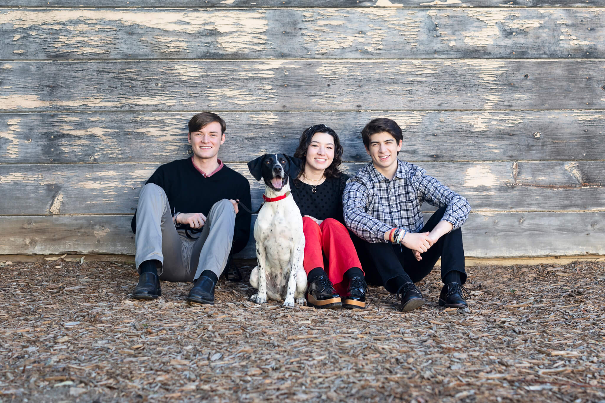 Family photo idea of three young adults on the ground in front of a wooden wall with a black and white dog between them. 