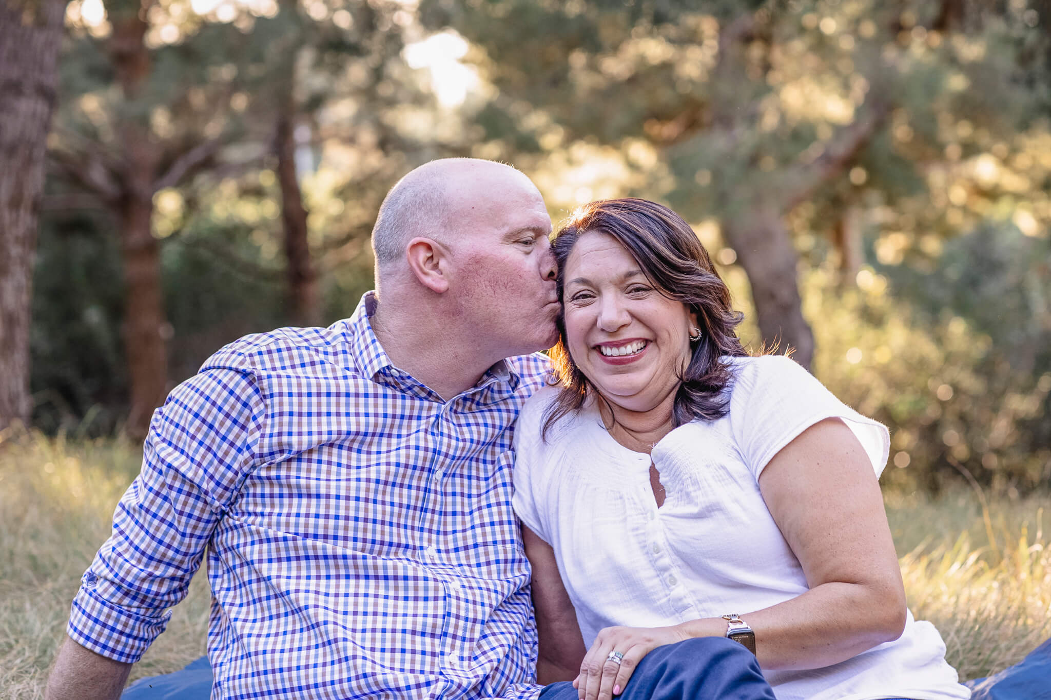 A man kisses a smiling woman on the cheek as they sit together outdoors on the grass.