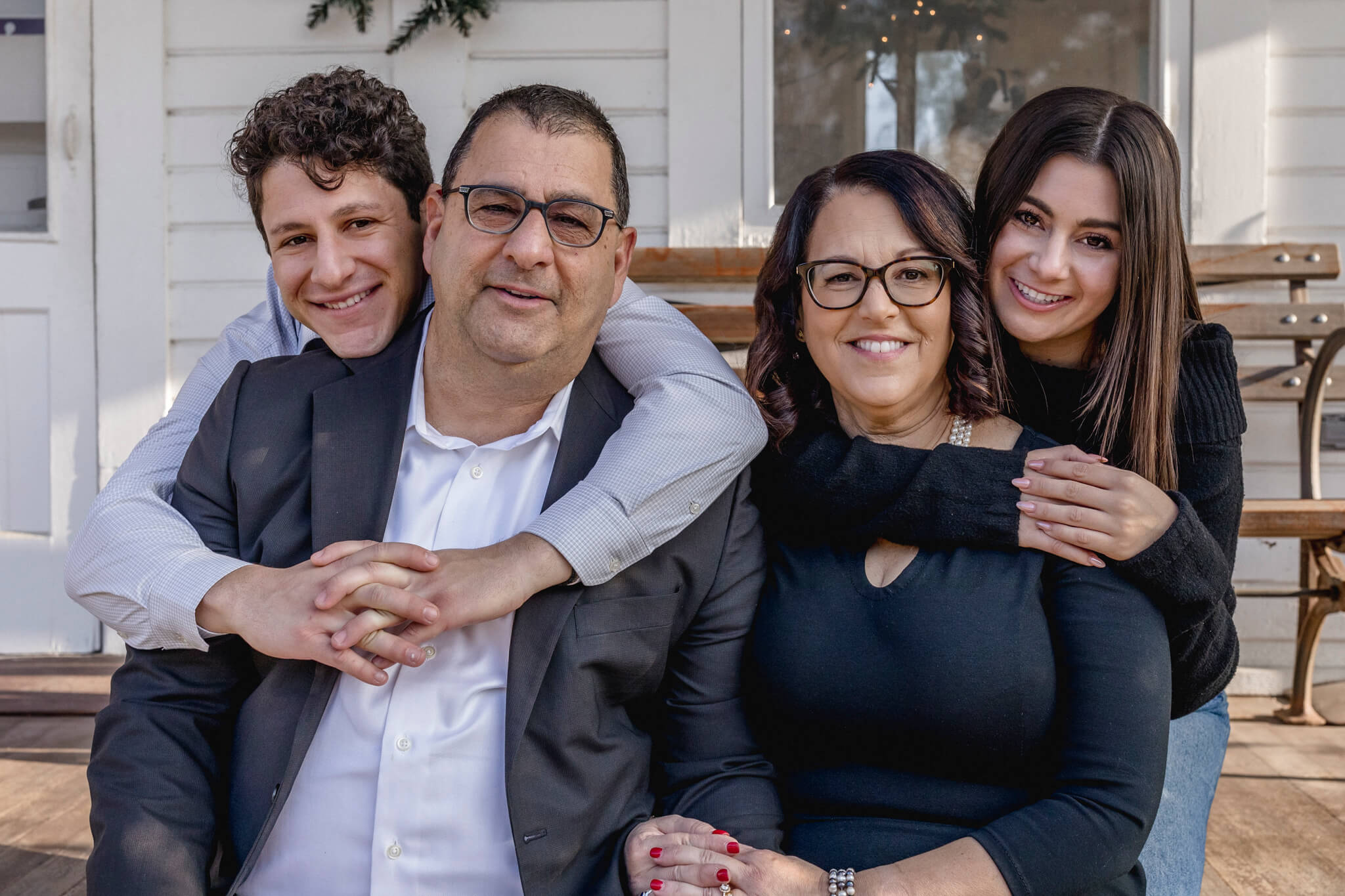 A family of four sits on a porch bench, smiling at the camera. The parents sit in front while their two teenage children stand behind them.