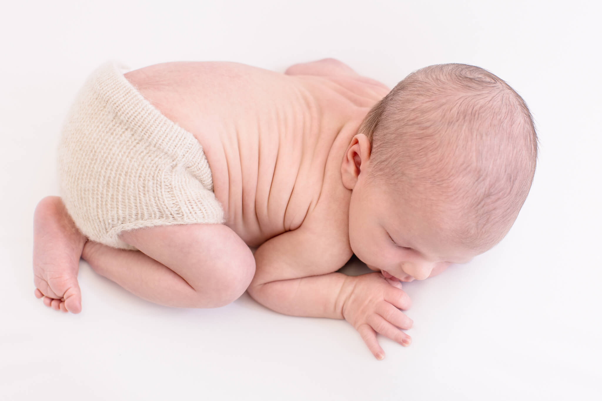 A newborn baby boy laying on his tummy with a knit diaper cover sleeping with his hand near his face on a soft white fabric.