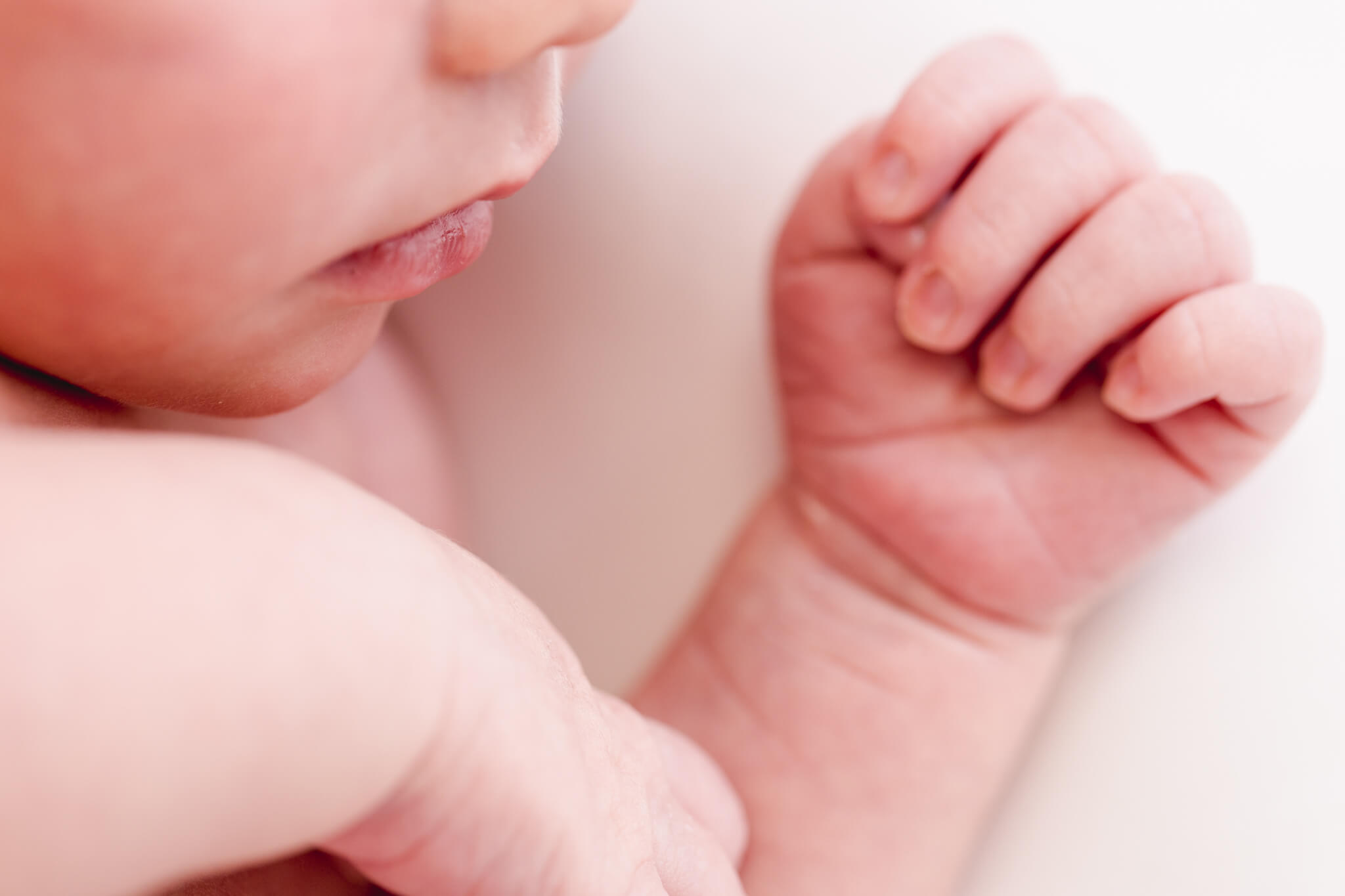 A sleeping newborn baby's lips and tiny hand with curled fingers resting on a soft white blanket.