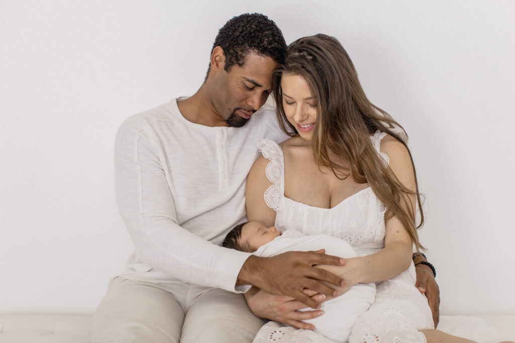 A couple in white clothing sits closely together looking down at their newborn baby at their Newport Beach newborn photoshoot