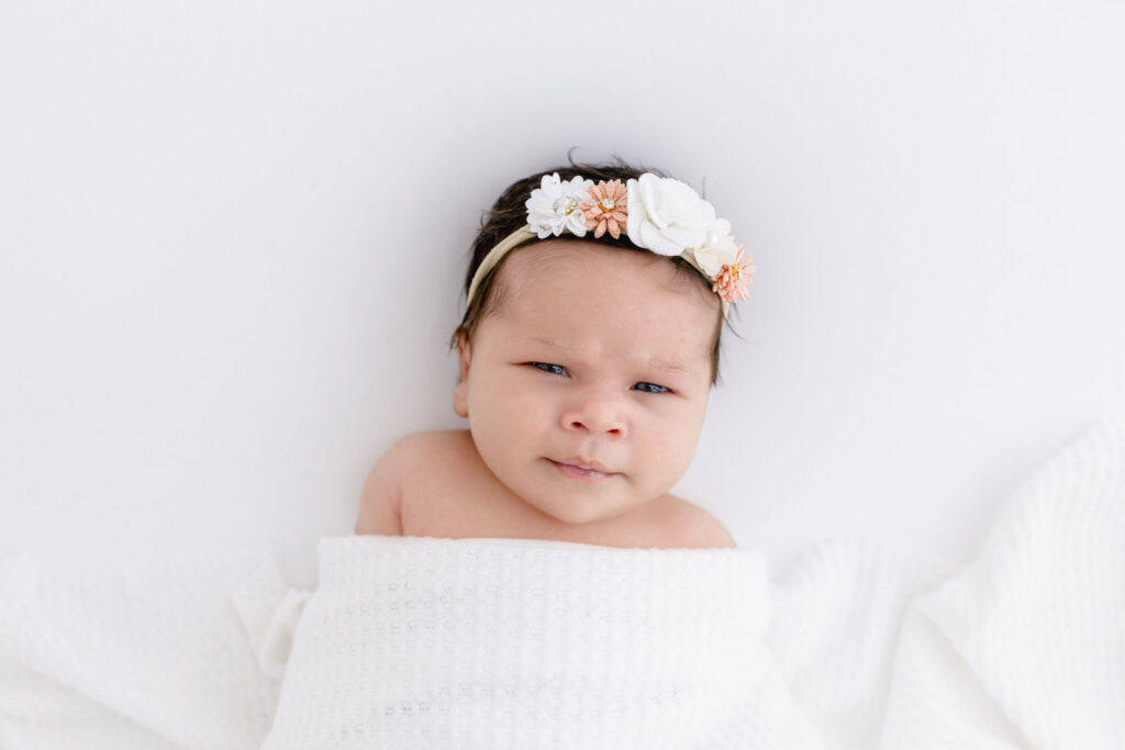 A baby with dark hair, wearing a floral headband, is wrapped in a white blanket.