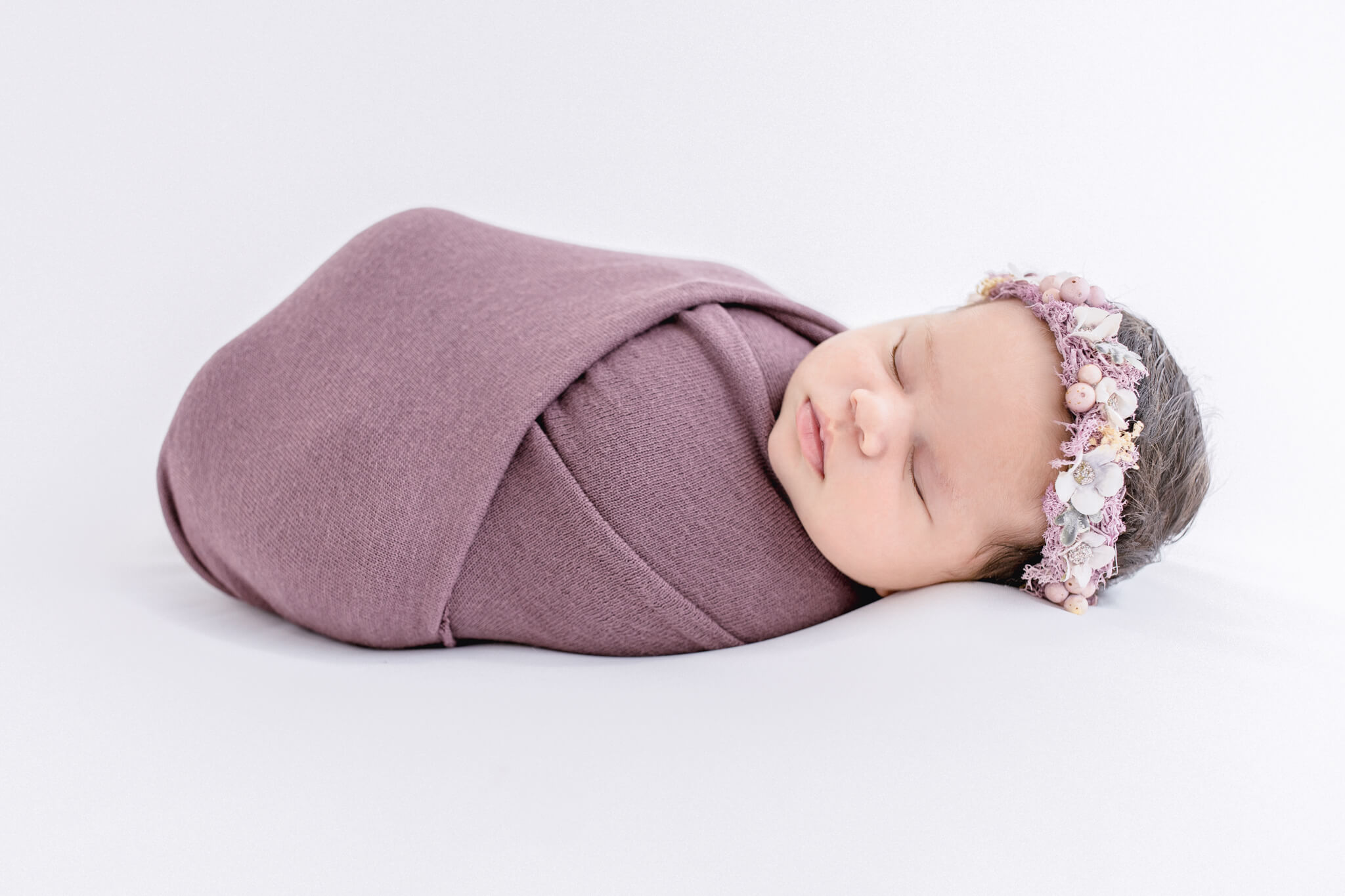 Three week old sleeping newborn baby girl swaddled in a purple wrap and floral headband laying down on a white fabric