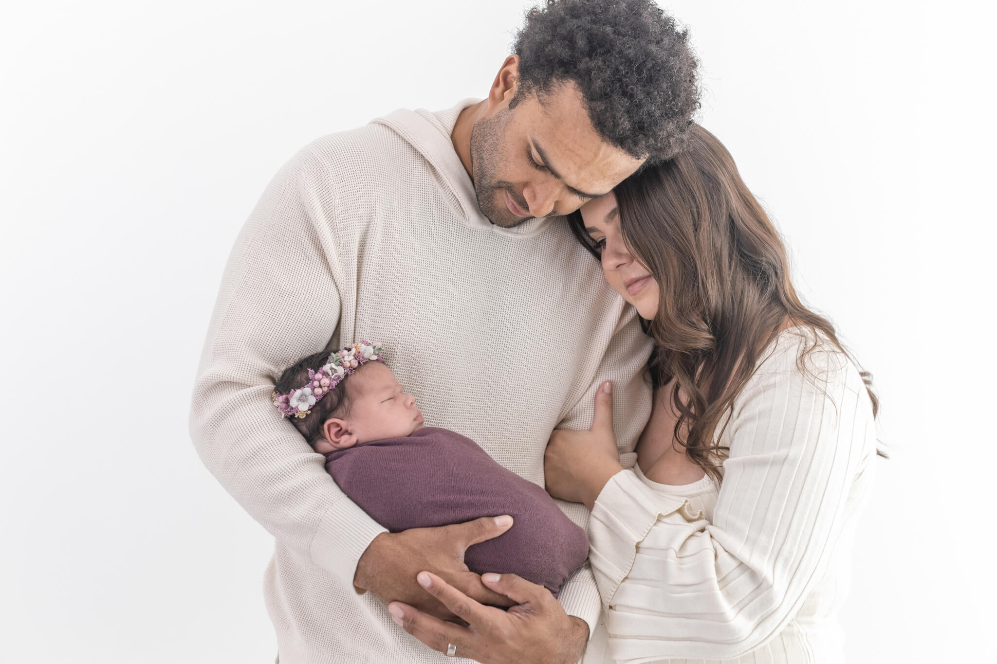 A couple embrace leaning their heads together while holding a baby wrapped in a purple blanket in Jennifer's Orange County studio.