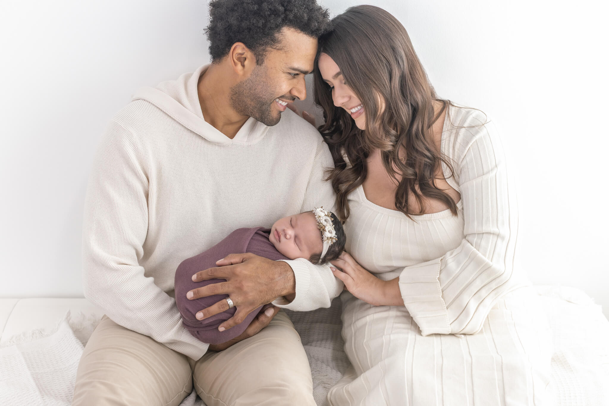 New parents holding a swaddled baby in a purple blanket and floral headband during an Orange County newborn studio photography session.