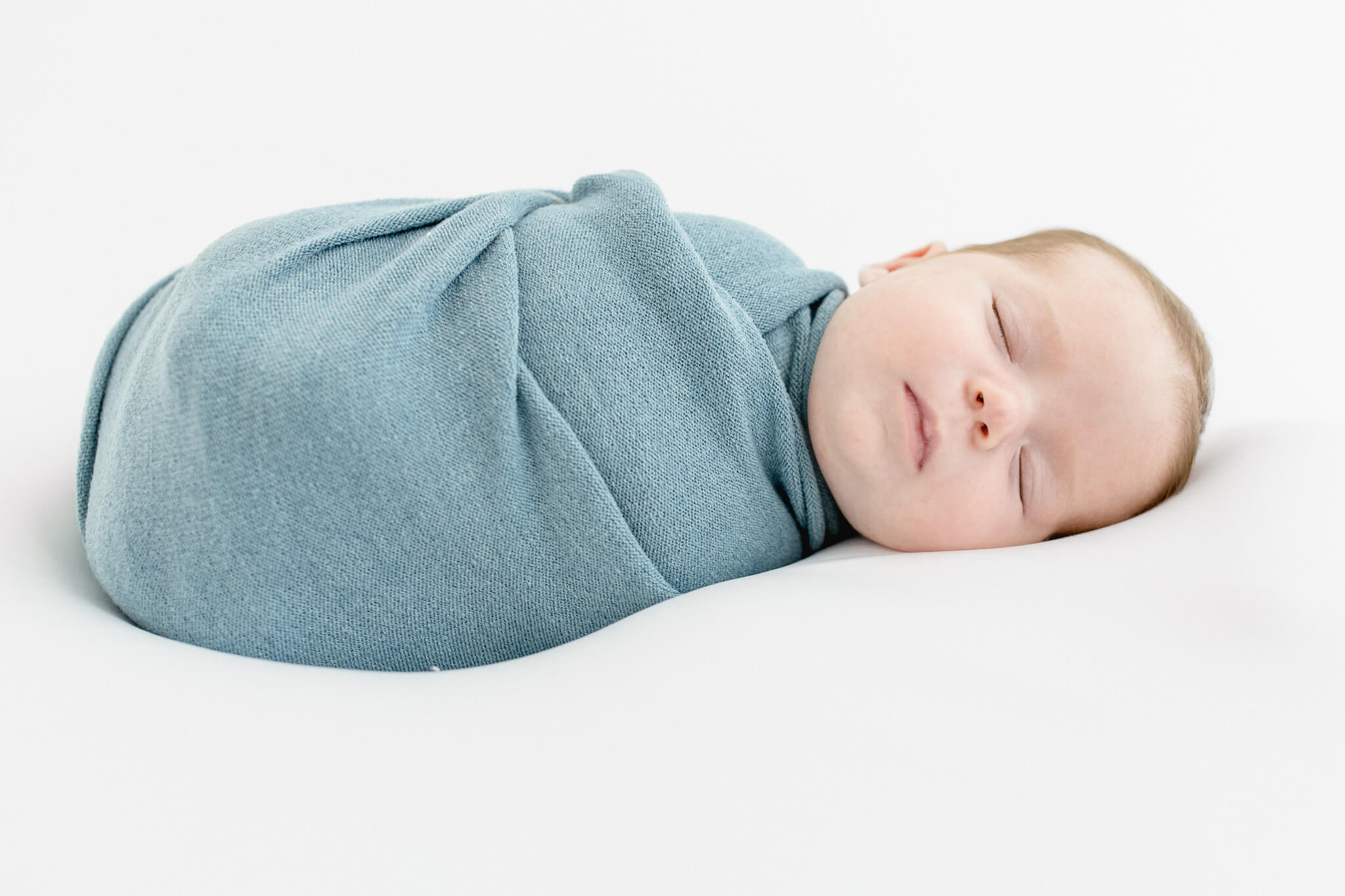 A sleeping baby girl swaddled snugly in a soft, light blue blanket, lying on a white surface with eyes closed and a peaceful expression.