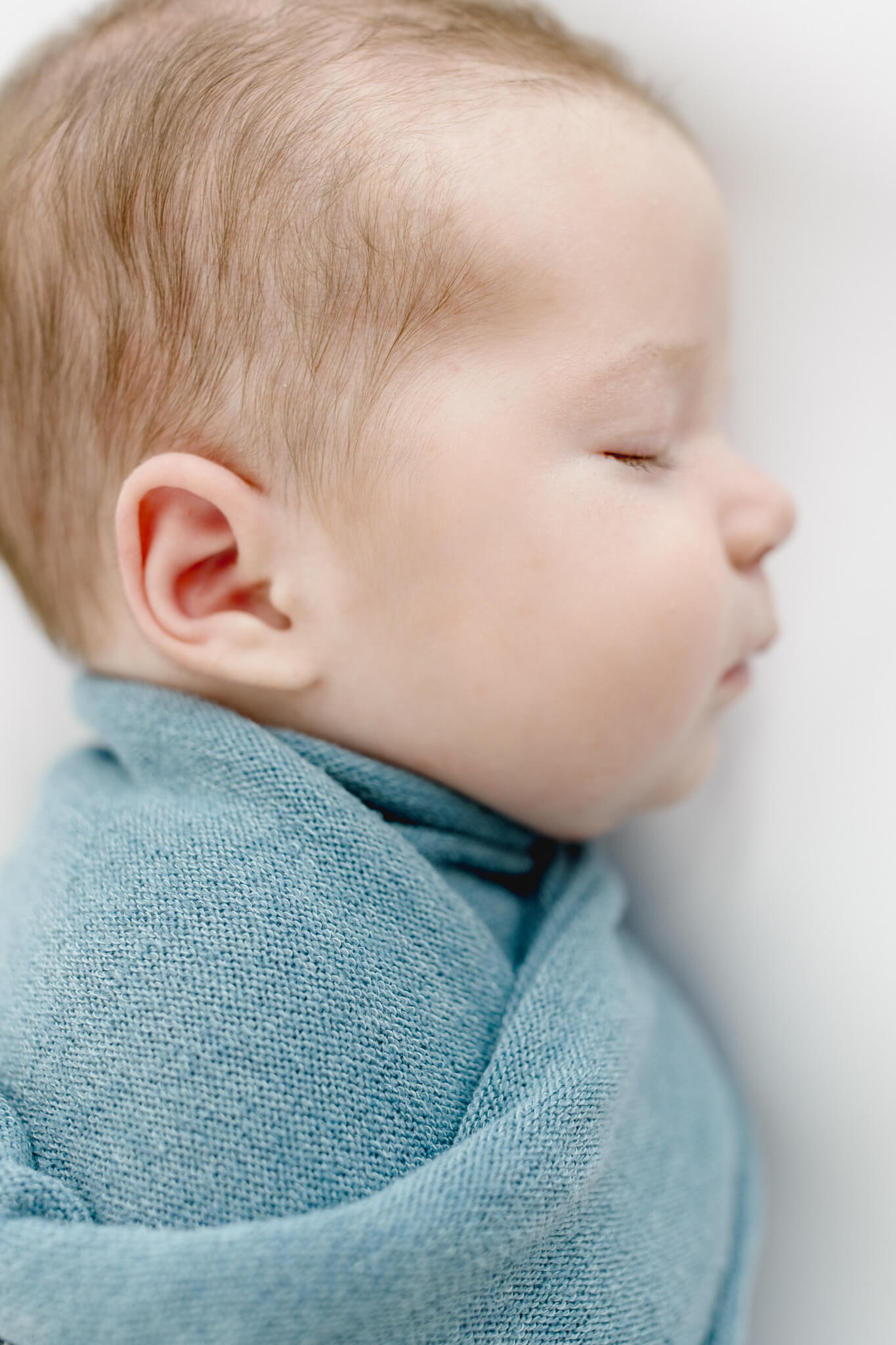 A sleeping newborn baby boy with light brown hair wrapped in a soft blue blanket lies on a white fabric background with eyes closed.