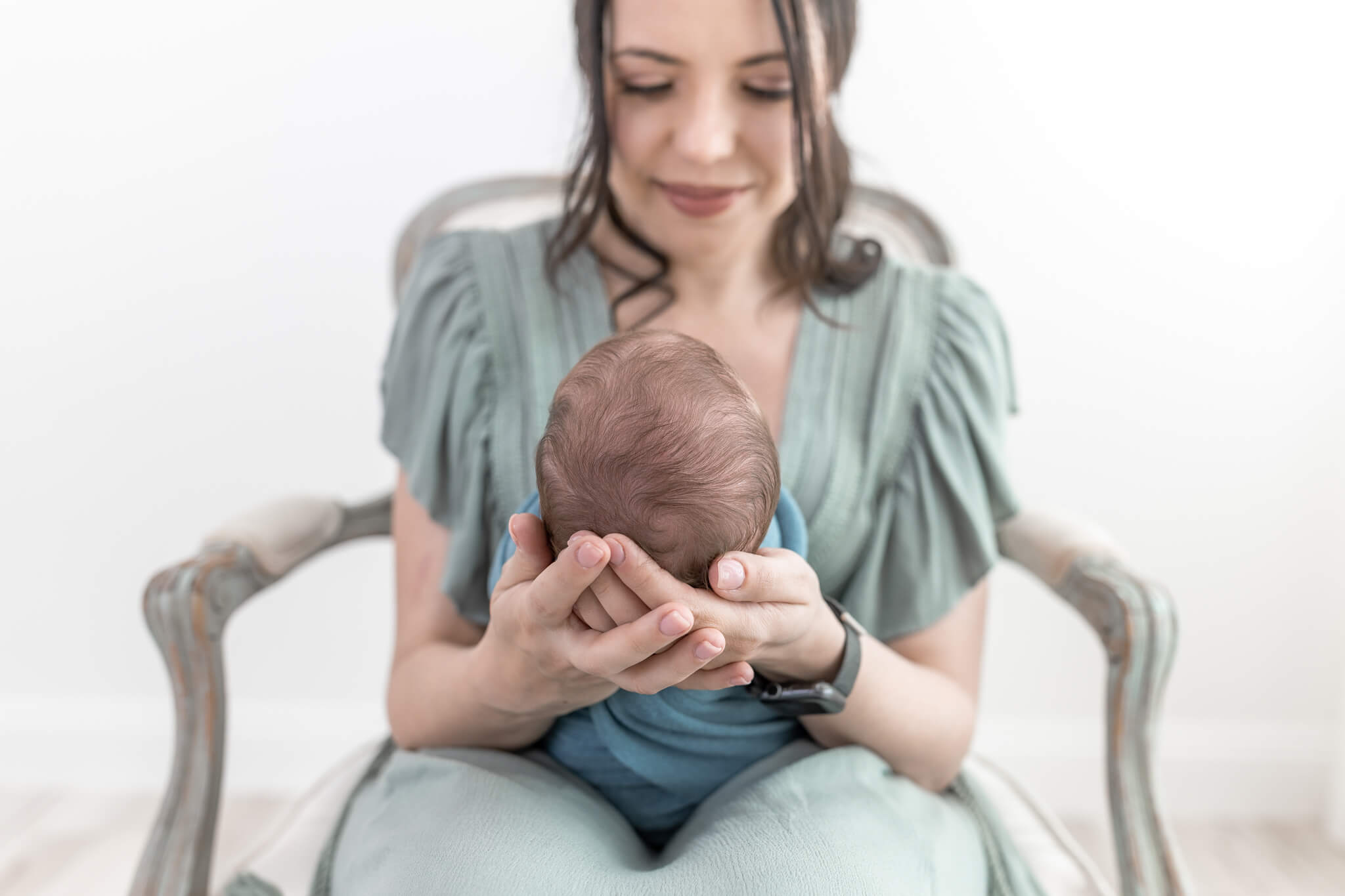 A mother in a sage green dress smiles down at her newborn wrapped in blue while gently holding his head in a vintage chair.