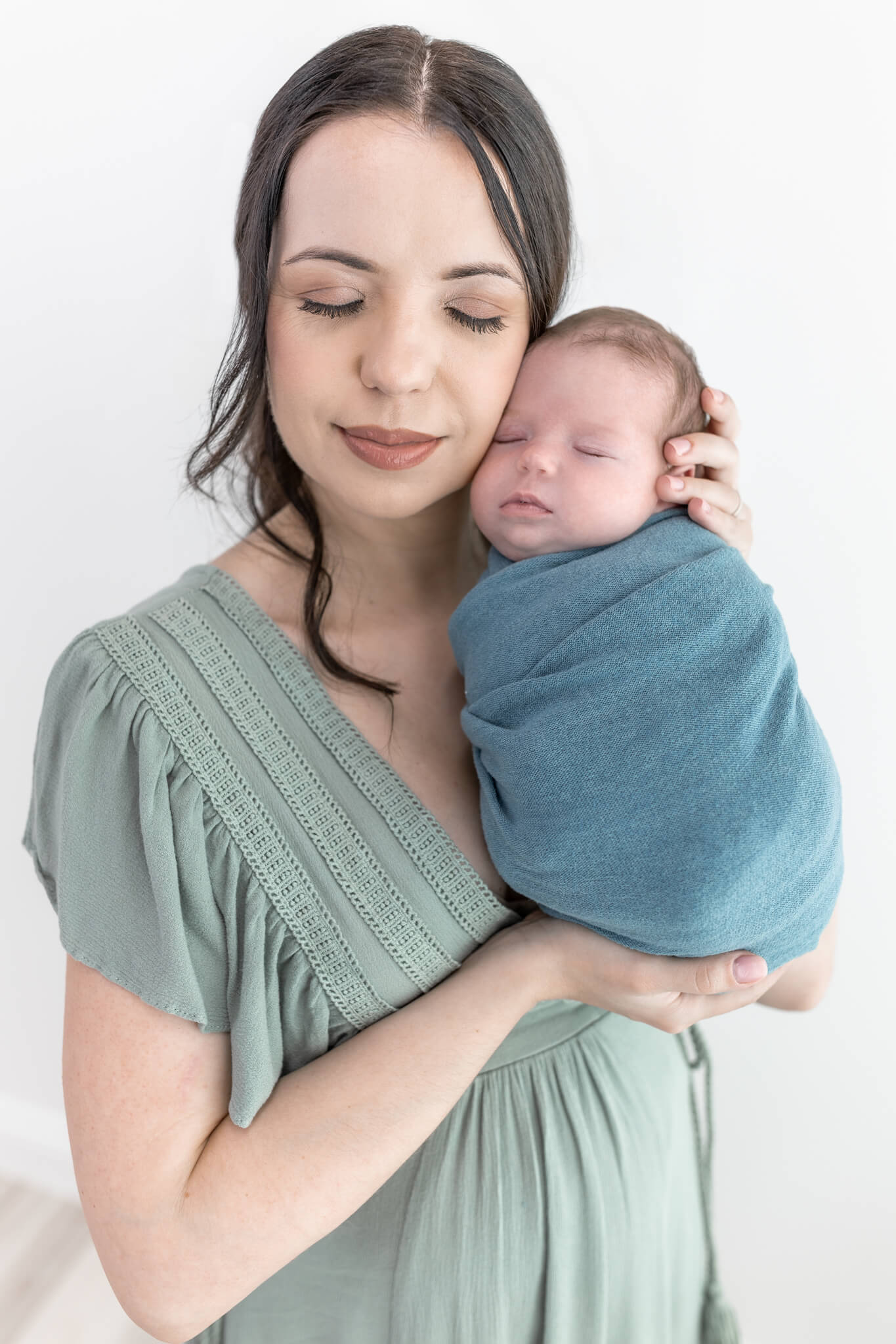 new mom in a sage green dress with her eyes closed and a peaceful expression holding her newborn son in a blue swaddle