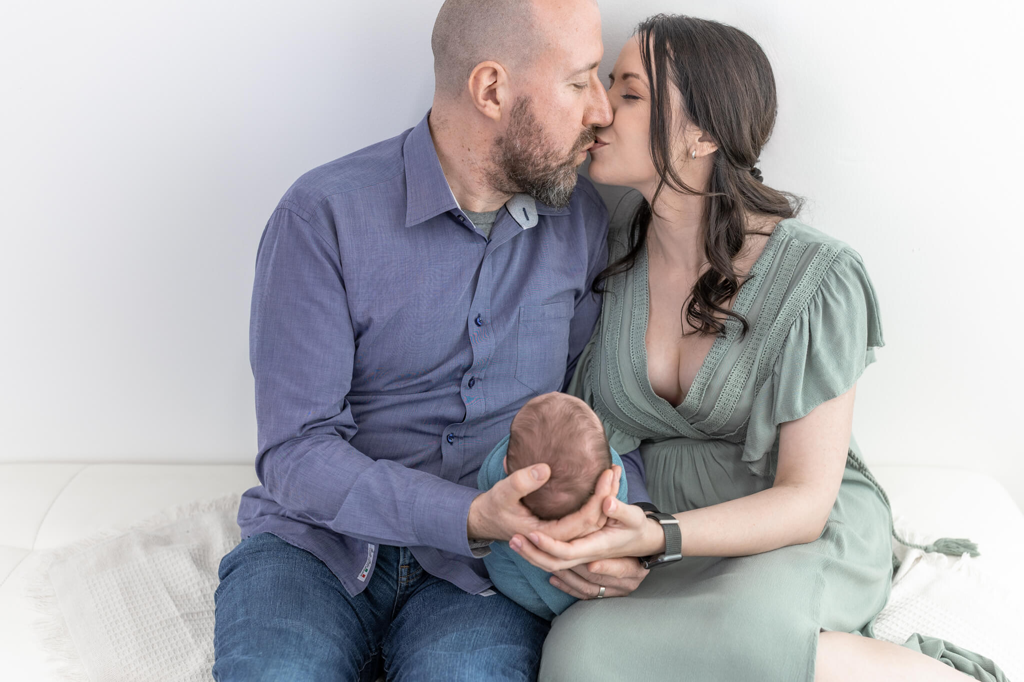 mom and dad both holding their newborn daughter in their lap as they tenderly kiss