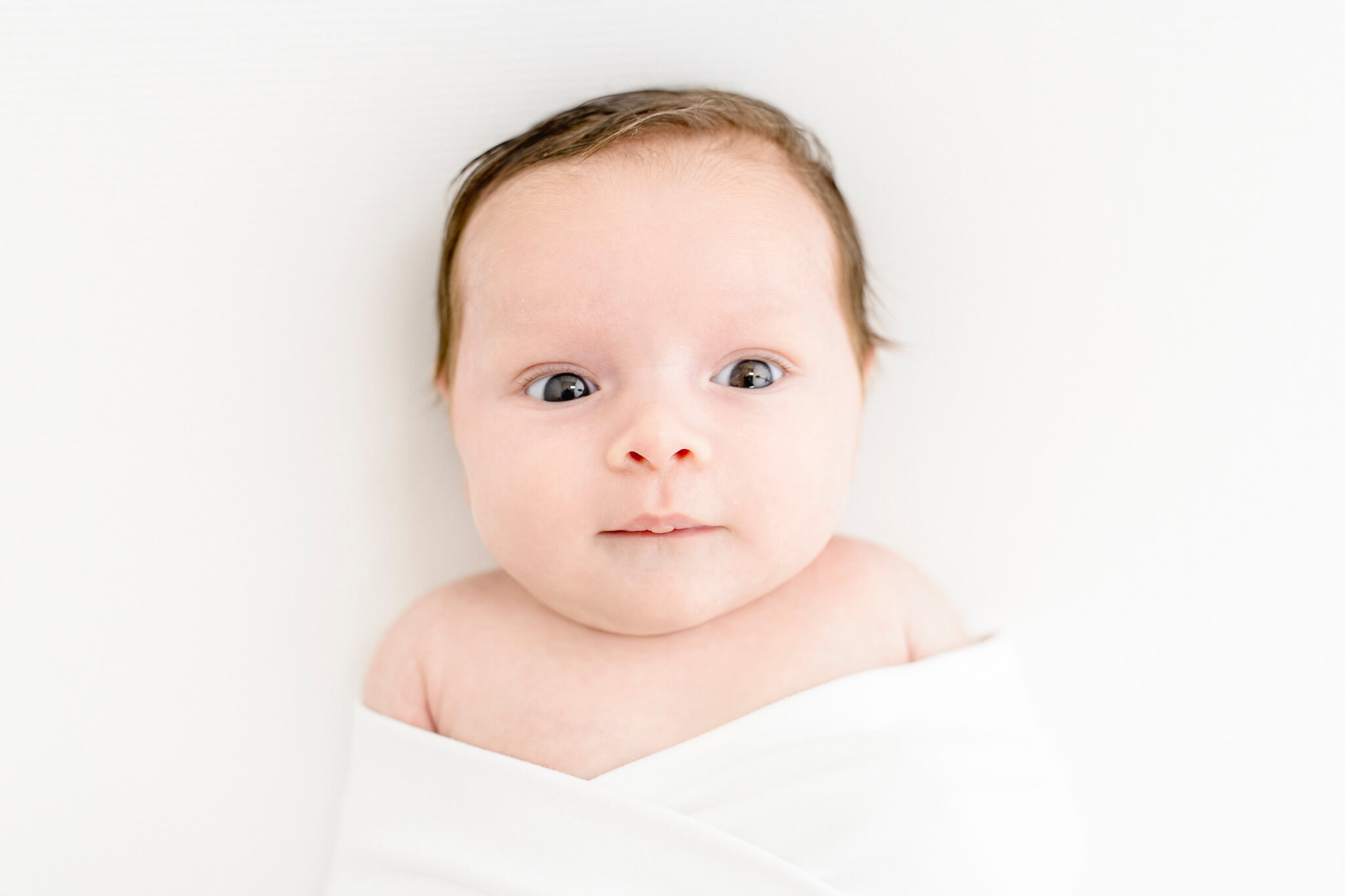 adorable 4 week old newborn wide awake with dark hair looking at the camera in a pure white swaddle laying on solid white fabric.