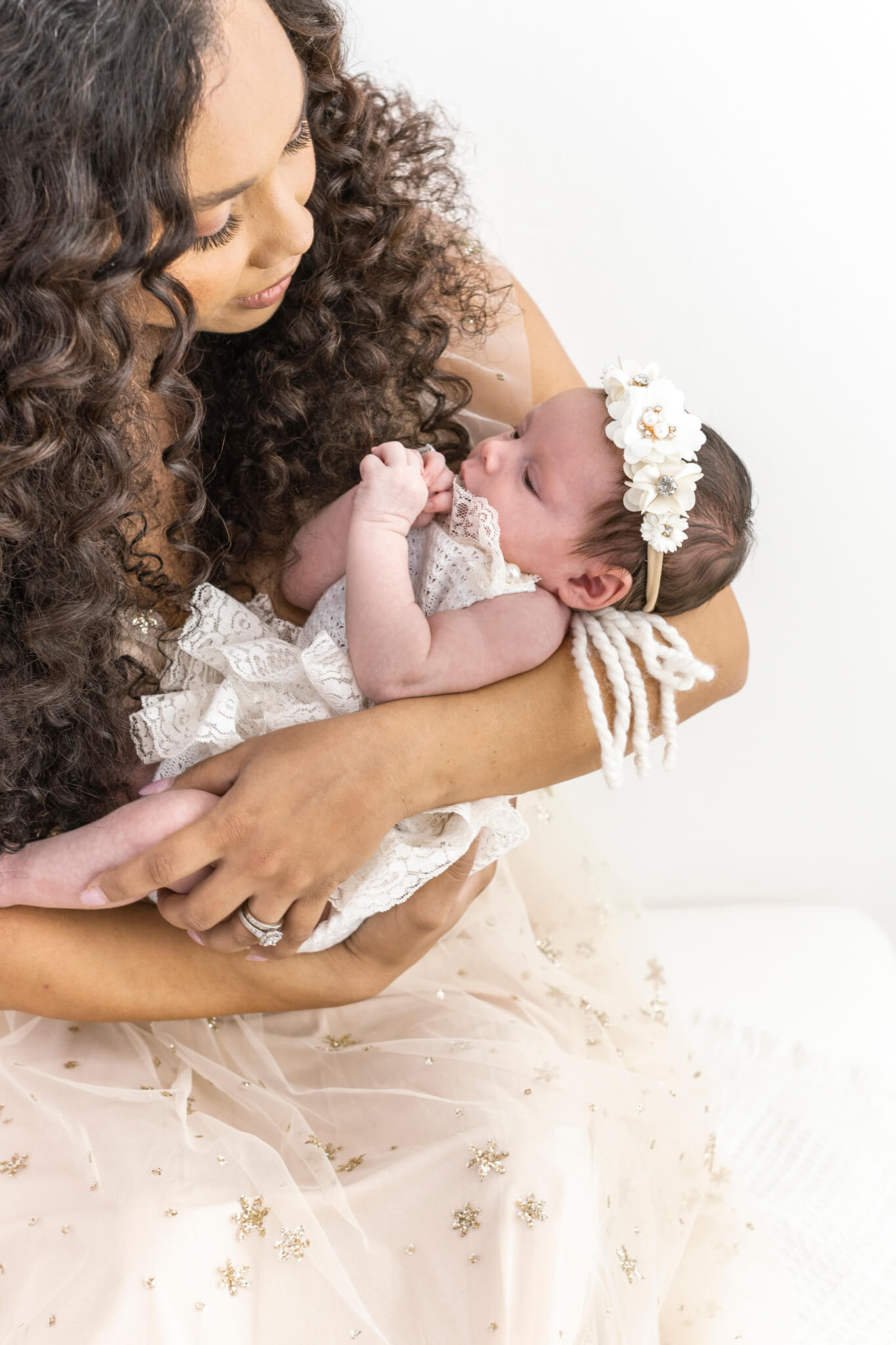 new mom in a tulle gown sitting on a settee looking down with love at her newborn baby girl in a white outfit and floral headband