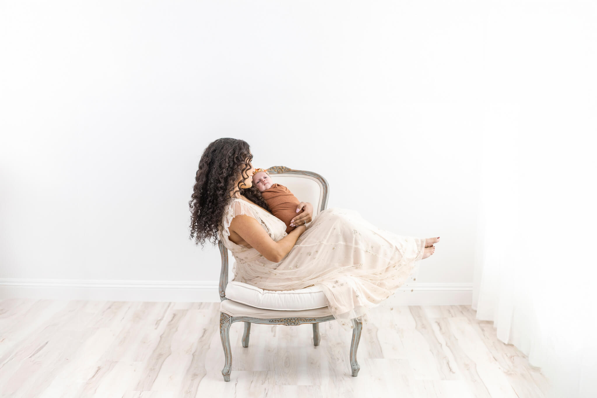 new mom with long dark hair in cream long dress sitting in vintage armchair holding her saddled newborn during an Orange County photoshoot.