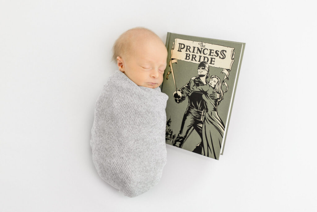 A newborn baby wrapped in gray sleeps next to a copy of The Princess Bride book.