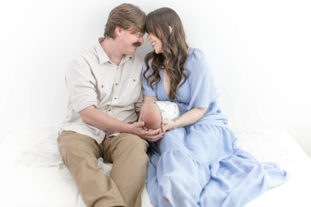 A man and woman sit closely together on a white bed smiling and touching foreheads while holding a newborn baby in Orange County.