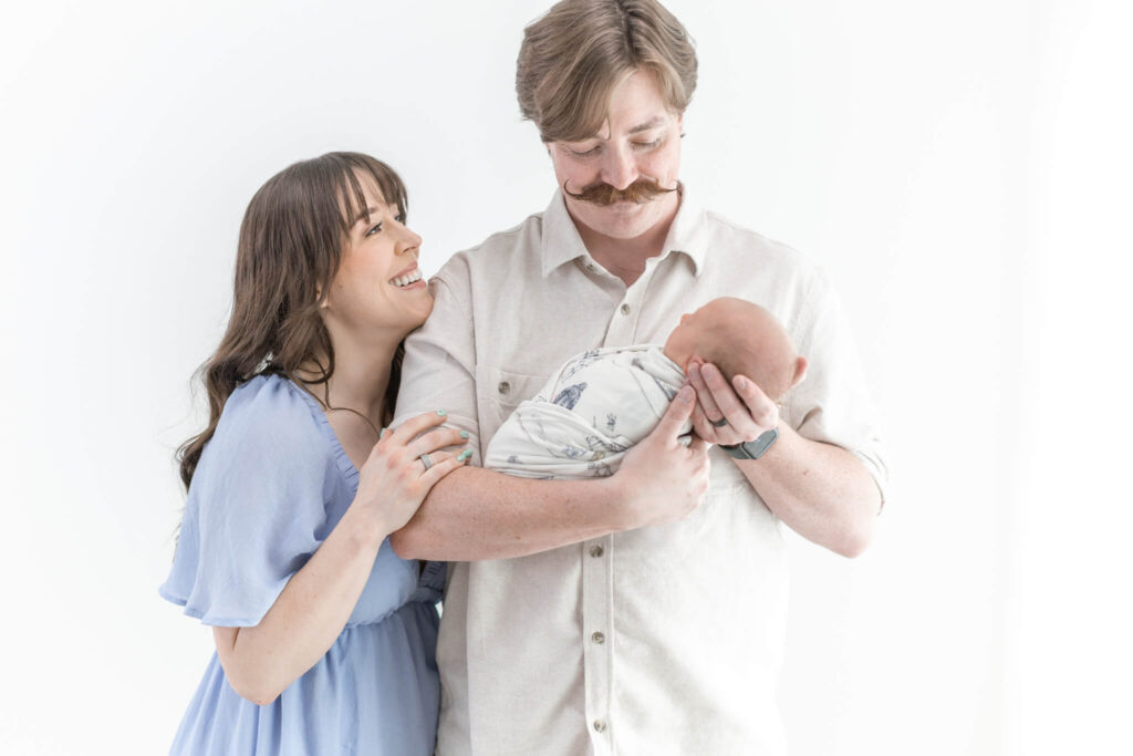 A smiling woman in a blue dress stands next to a man holding a swaddled newborn baby.