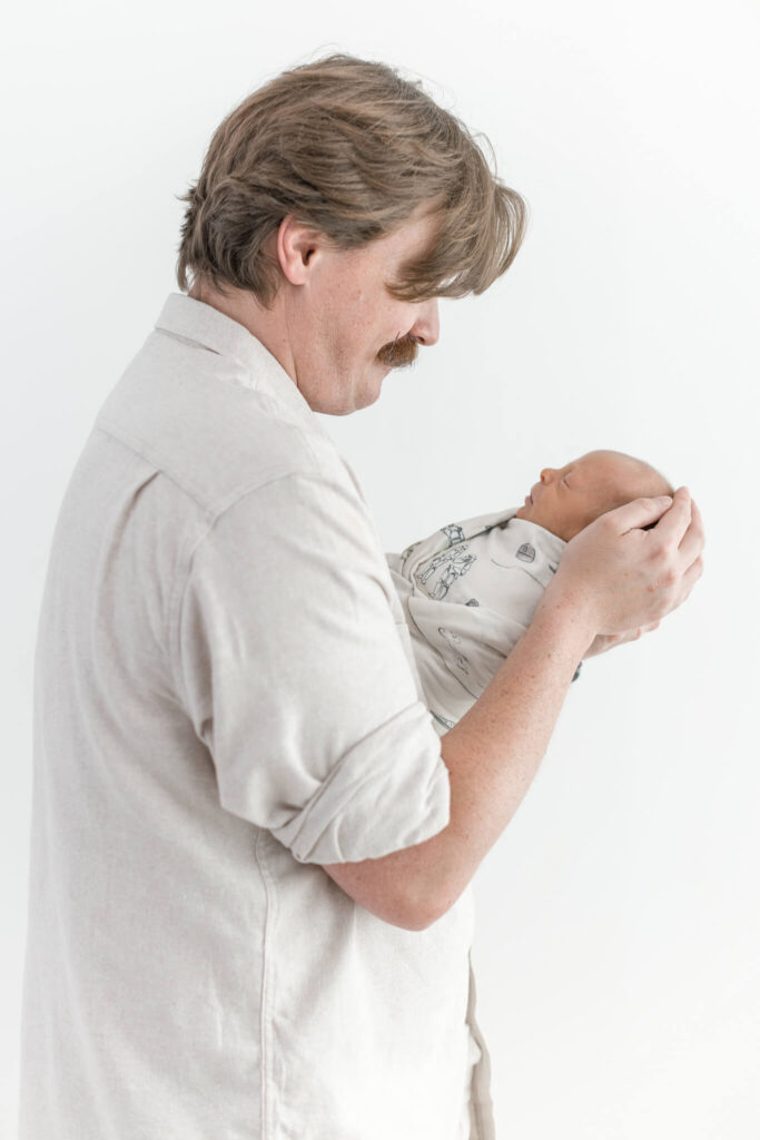 A man holds a swaddled newborn baby looking down smiling at Jennifer Imus Photography's newborn studio.