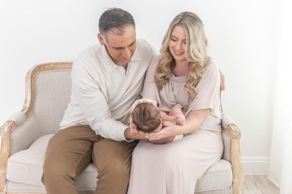 A man and woman sit on a light-colored bench, holding a newborn baby at an Orange County photo studio.